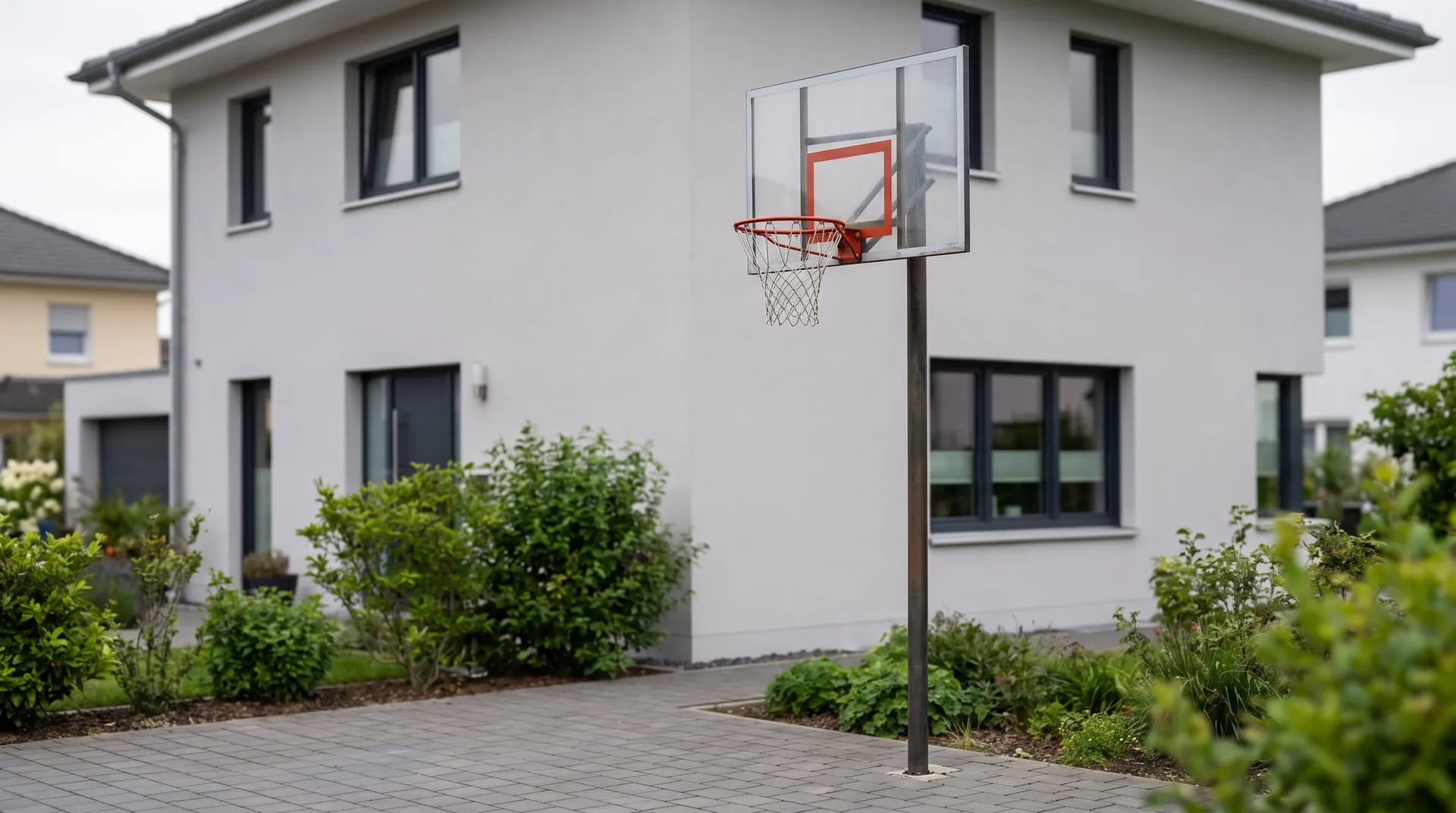 Basketballkorb auf einem eigenen Stahlpfosten auf der gepflasterten Einfahrt, mit Abstand vor einer glatt verputzten, gedämmten Hausfassade